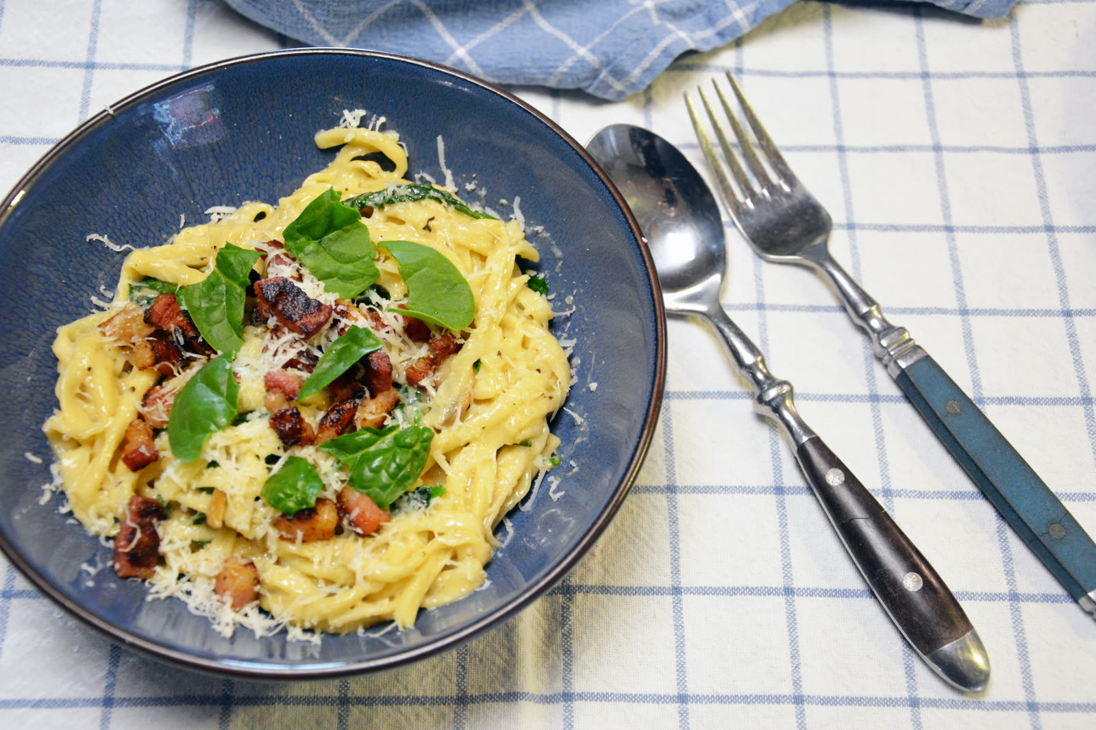 Pasta con bacon, espinacas y parmesano rallado en un bol azul sobre un paño de cocina de cuadros