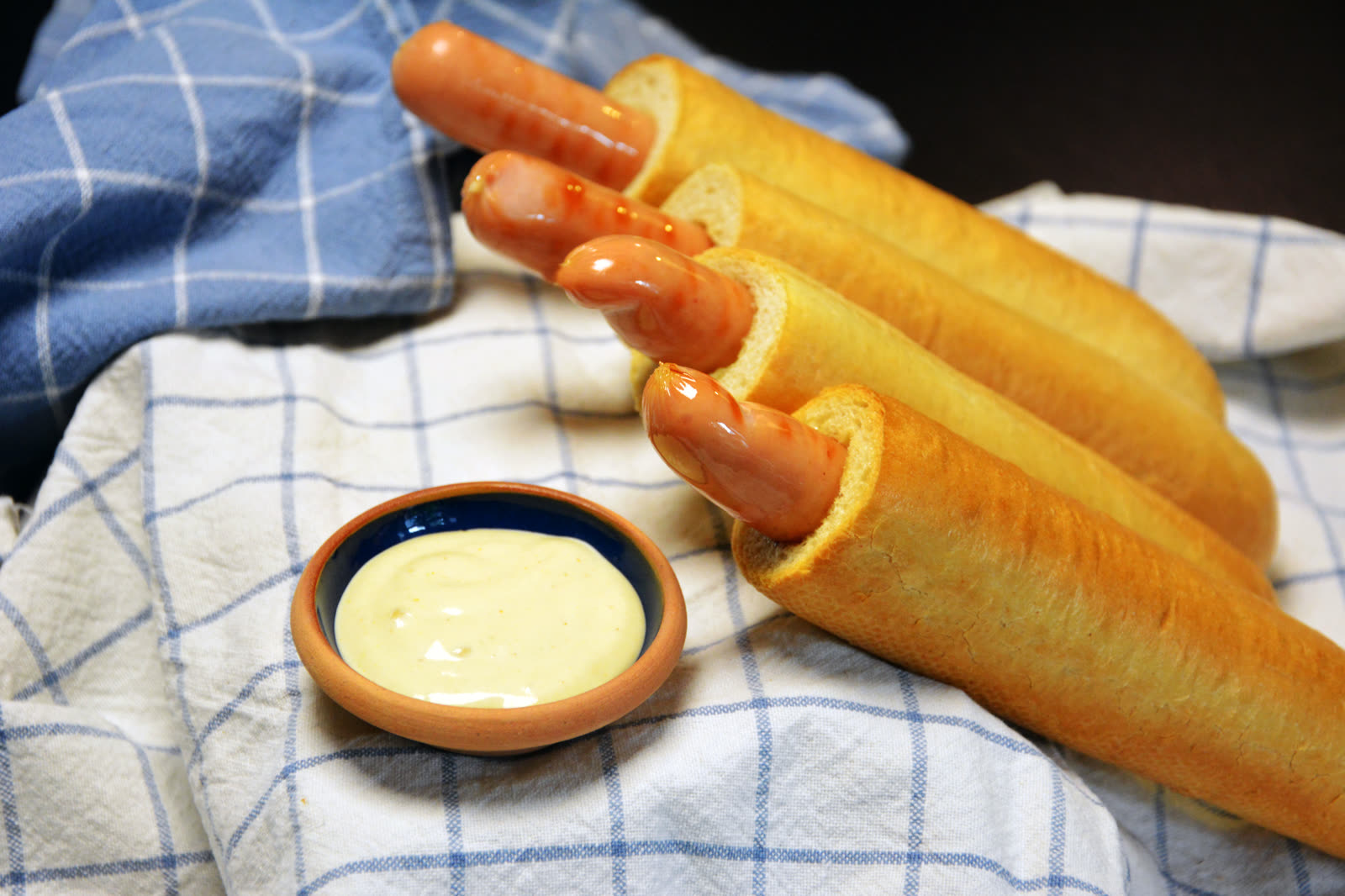 Cuatro perritos calientes franceses con la salchicha dentro del agujero sobre un paño de cuadros azules junto a un cuenco con salsa clara
