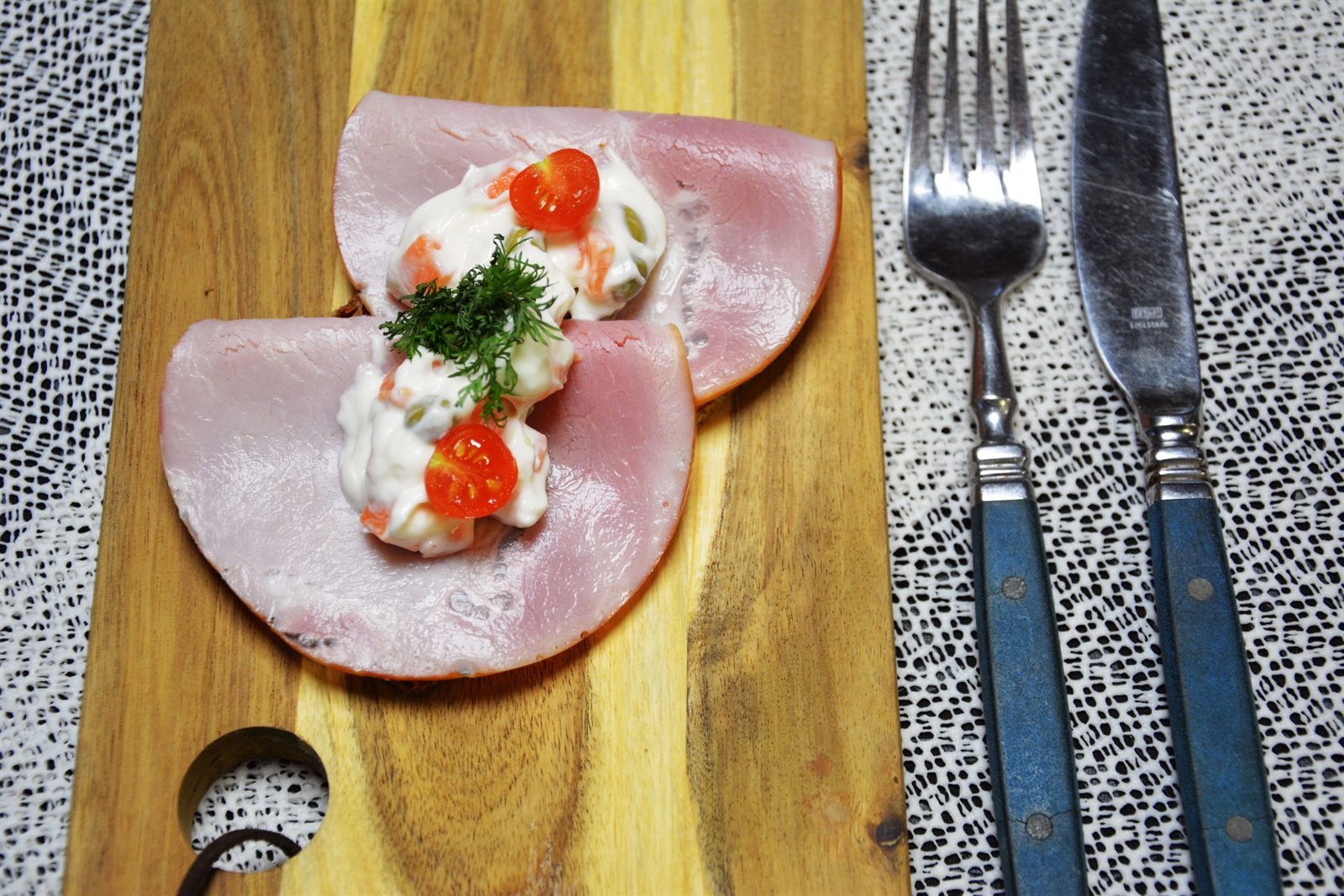 Ensaladilla sin patata con dados de zanahoria y guisantes sobre dos lonchas de jamón cocido dobladas encima de pan de centeno, con medio tomate cherry y eneldo