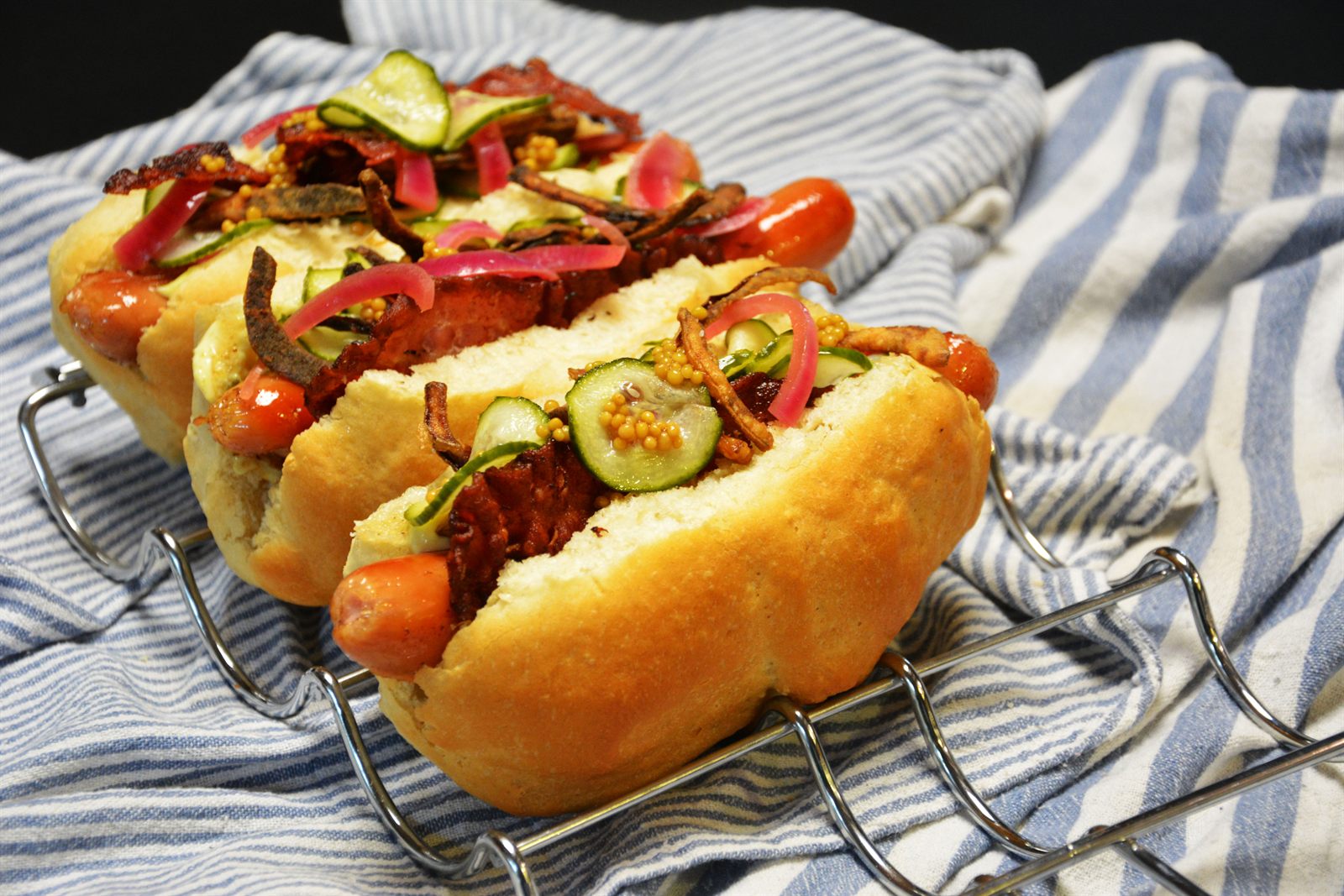 Perrito caliente danés con bacon, cebolla roja encurtida, cebolla crujiente, ensalada de pepino y granos de mostaza encurtidos